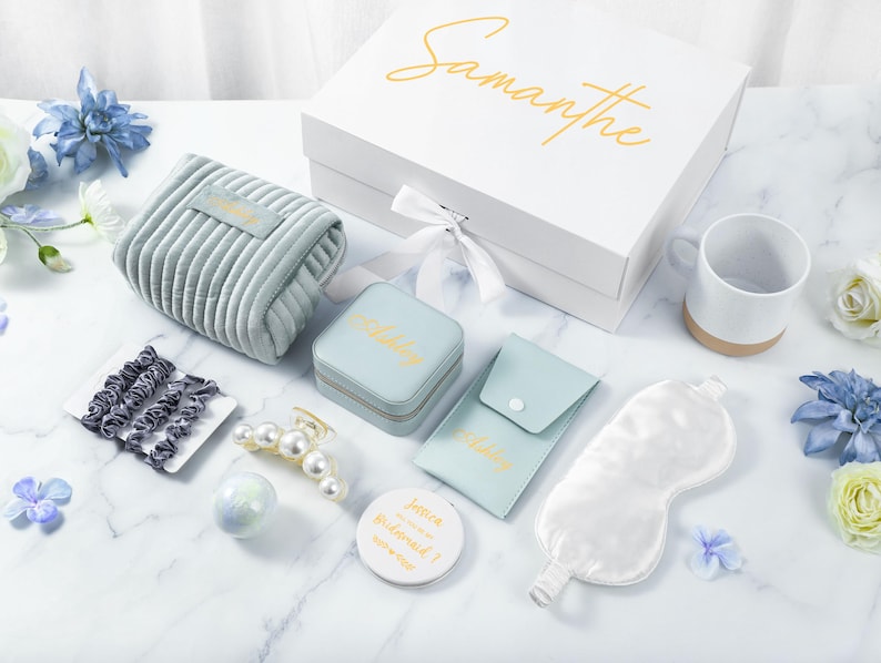 May include: A gift box with the name Samantha on the lid. The box contains a variety of items, including a blue velvet pouch, a small jewelry box, a sleep mask, a mug, hair clips, and a bath bomb. The box is perfect for a bridesmaid gift.