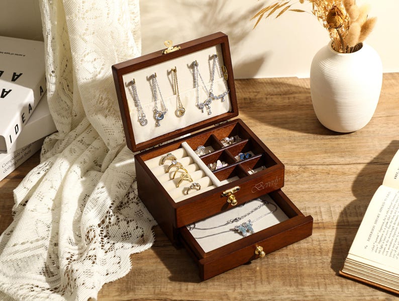 May include: A wooden jewellery box with two drawers. The top drawer has compartments for rings, earrings, and necklaces. The bottom drawer is open and shows a necklace inside. The box is brown with gold accents.