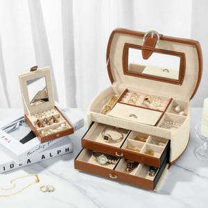 Könnte beinhalten: Eine braune Leder-Schmuckschatulle mit einem Spiegel und mehreren Fächern zum Verstauen von Ringen, Ohrringen, Halsketten und Uhren. Die Schatulle hat zwei Schubladen und ein oberes Fach mit einem Klappdeckel.