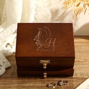 Puede incluir: Una caja de madera para joyas con tapa abatible y dos cajones. La tapa está grabada con un diseño de mariposa y el texto "Hannah Ali".