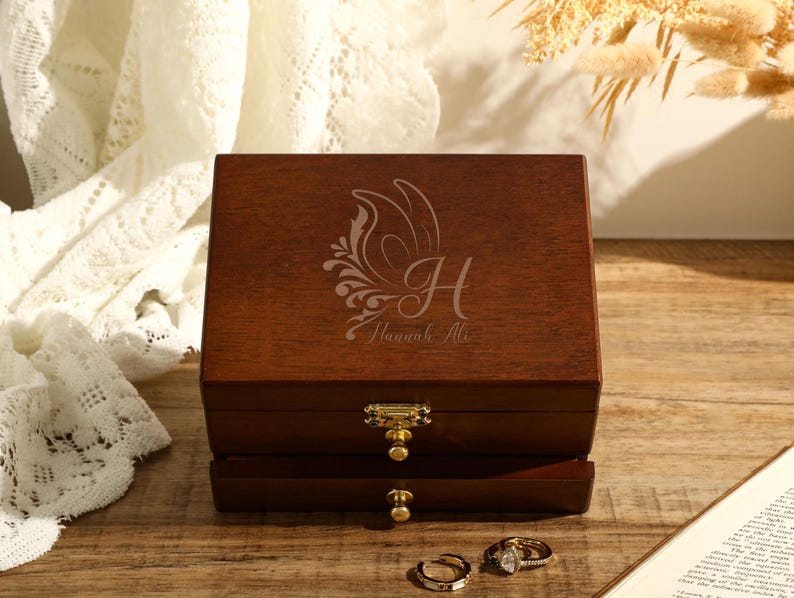 May include: A wooden jewellery box with a hinged lid and two drawers. The lid is engraved with a butterfly design and the text "Hannah Ali".