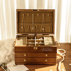 May include: A wooden jewelry box with multiple compartments and drawers, open to display necklaces and rings. The box is brown with gold hardware and a light-colored interior. The text "You are the one I have been looking for" is printed on the front.