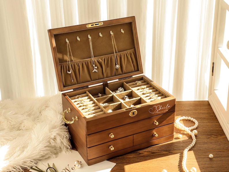 May include: A wooden jewelry box with multiple compartments and drawers. The box is open, revealing necklaces hanging from the lid and rings in the compartments. The box has a gold-colored handle and drawer pulls. A strand of pearls is draped nearby.