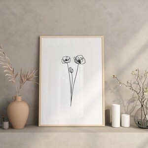 May include: A framed black and white line drawing of three poppies on a white background. The frame is wooden and has a light brown finish. The drawing is displayed on a shelf with two vases and a plant.