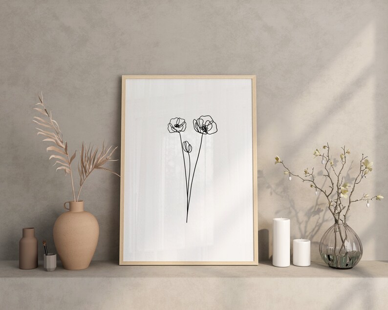 May include: A framed black and white line drawing of three poppies on a white background. The frame is wooden and has a light brown finish. The drawing is displayed on a shelf with two vases and a plant.