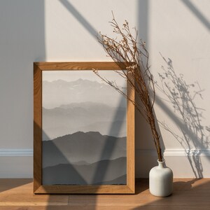 May include: A framed print of a black and white mountain landscape with a dried flower arrangement in a white vase in the foreground.