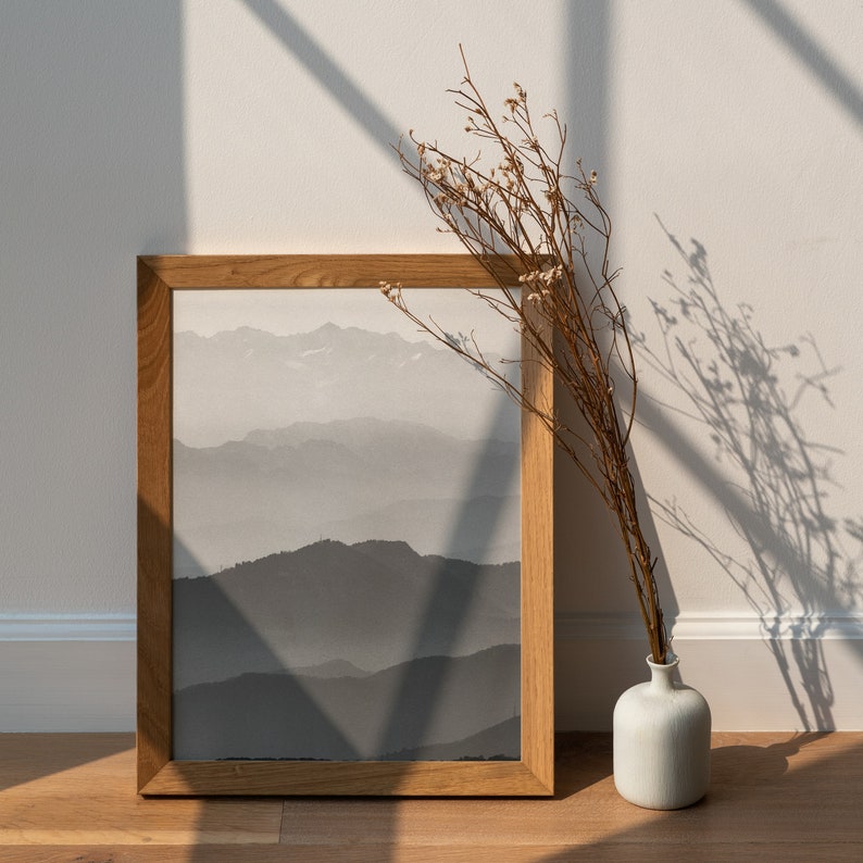 May include: A framed print of a black and white mountain landscape with a dried flower arrangement in a white vase in the foreground.