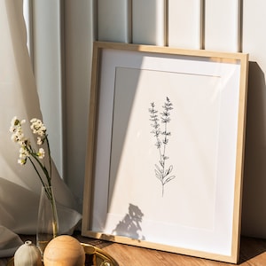 May include: A framed print of a simple line drawing of a sprig of lavender, with a wooden frame and a white mat. The print is sitting on a wooden floor with a gold tray and decorative objects.