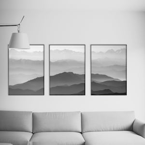 May include: Three black framed photographs of a mountain range with a hazy, layered effect in shades of gray. The photographs are hung on a white wall above a gray sectional sofa.