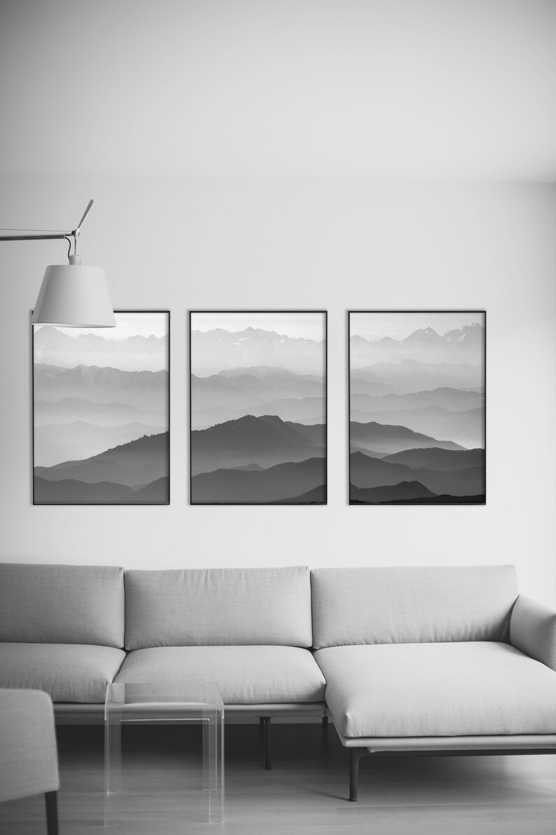 May include: Three black framed photographs of a mountain range with a hazy, layered effect in shades of gray. The photographs are hung on a white wall above a gray sectional sofa.