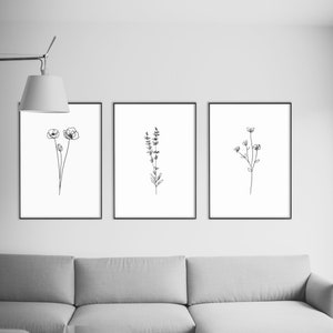 May include: Three black framed prints of line drawings of flowers and plants hanging on a white wall above a gray sectional sofa and a clear glass coffee table.