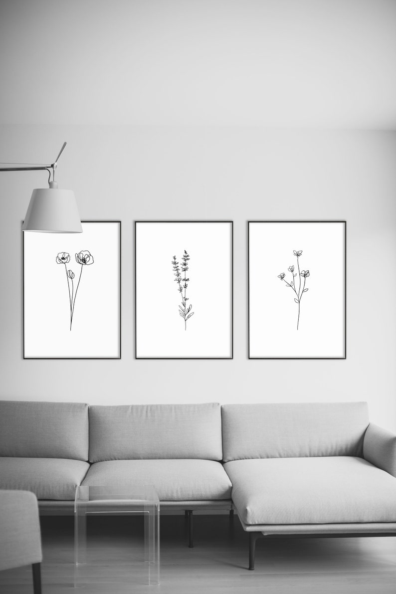 May include: Three black framed prints of line drawings of flowers and plants hanging on a white wall above a gray sectional sofa and a clear glass coffee table.
