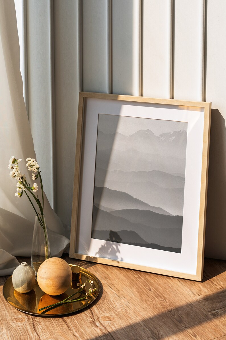 May include: A framed black and white photograph of a mountain range with a wooden frame. The photo is on a wooden floor with a gold tray, a small white vase, and a wooden ball.