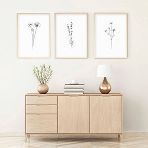 May include: Three black and white line drawings of flowers in simple frames on a white wall above a light wood dresser with two drawers and two doors. A small potted plant sits on the dresser.