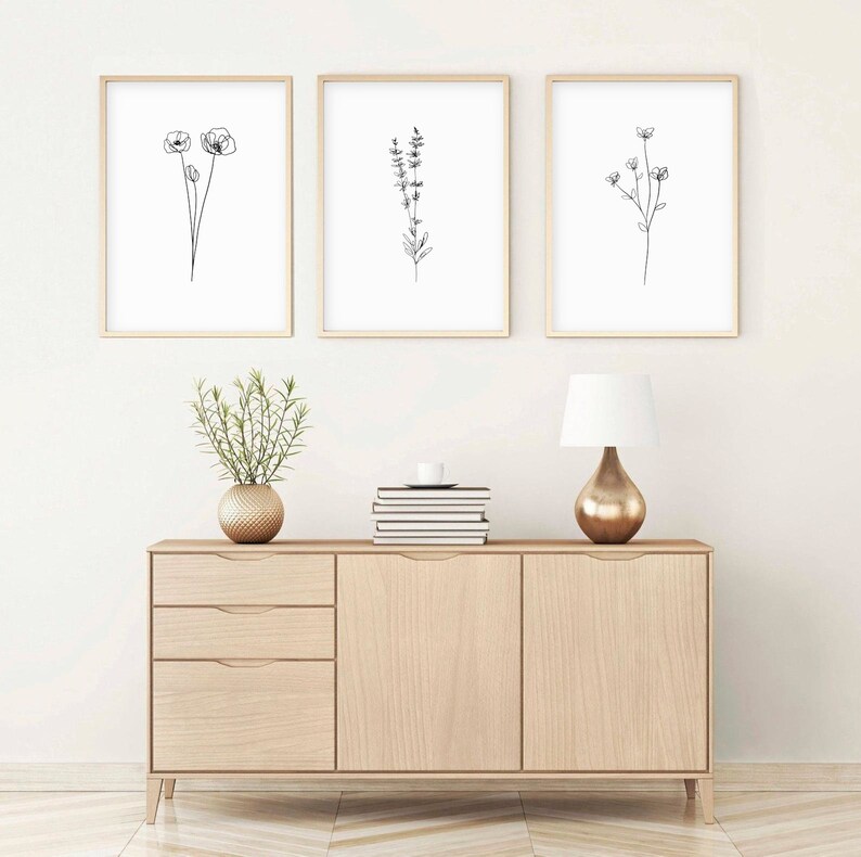 May include: Three black and white line drawings of flowers in simple frames on a white wall above a light wood dresser with two drawers and two doors. A small potted plant sits on the dresser.