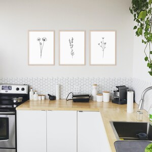 May include: Three black and white line drawings of flowers in simple frames, hung on a white wall above a kitchen counter with white cabinets and a stainless steel stove.