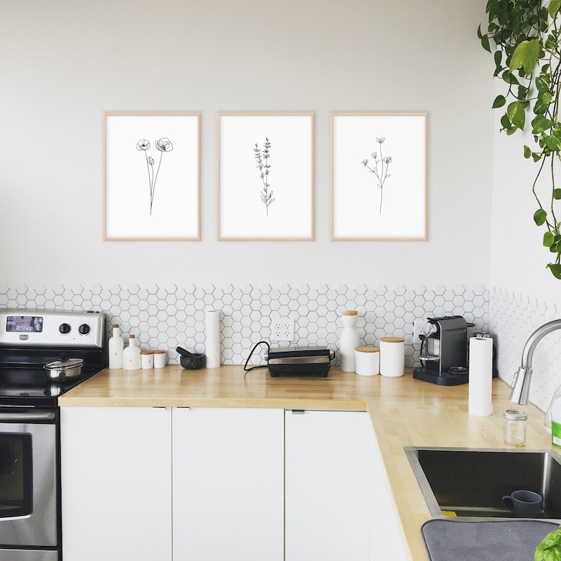 May include: Three black and white line drawings of flowers in simple frames, hung on a white wall above a kitchen counter with white cabinets and a stainless steel stove.