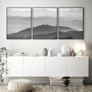 May include: Three black and white framed prints of a mountain range with fog in the background. The prints are hanging on a white wall above a white cabinet with a white ottoman in front of it.