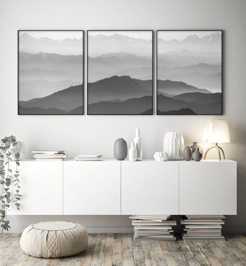 May include: Three black and white framed prints of a mountain range with fog in the background. The prints are hanging on a white wall above a white cabinet with a white ottoman in front of it.