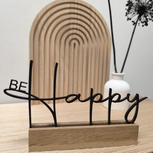 Könnte beinhalten: Ein schwarzes "BE Happy"-Schild auf einem Holzsockel. Die Schrift ist verbunden. Im Hintergrund befindet sich eine hölzerne Wanddekoration in Bogenform und eine weiße Vase mit schwarzen Stielen.