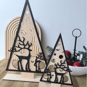 Könnte beinhalten: Zwei Weihnachtsbaumdekorationen aus Holz. Die Bäume sind dreieckig mit schwarzem Rand und natürlichem Holzinnenraum. Jeder Baum hat ausgeschnittene Kreise und schwarze Silhouetten von Rentieren. Die Bäume stehen auf Holzsockeln.