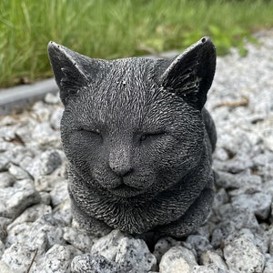 Puede incluir: Una estatua de gato de hormig&oacute;n gris con los ojos cerrados, tumbada en un lecho de peque&ntilde;as piedras grises.