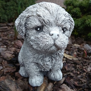 May include: A small, gray, concrete statue of a dog with a fluffy, textured coat. The dog is sitting with its head tilted down and its eyes closed.