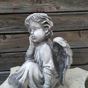 May include: A gray stone sculpture of a cherub angel with wings, sitting with its head resting on its hand. The angel is wearing a long robe and has a sweet expression on its face.