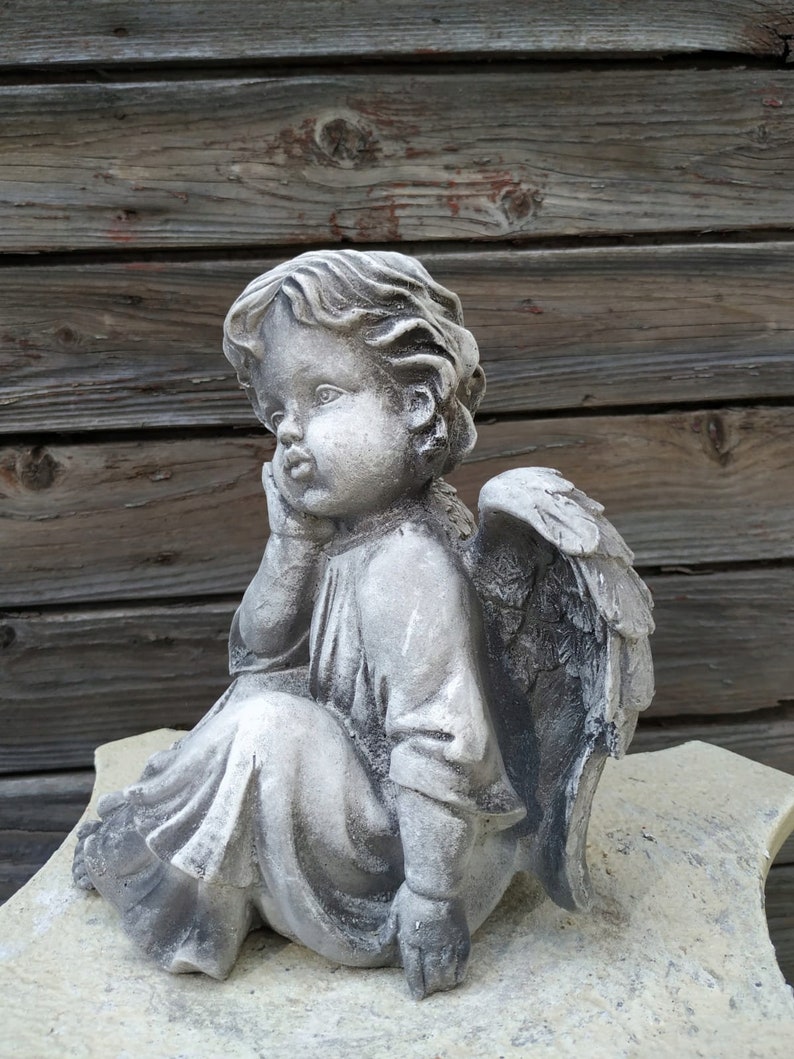 May include: A gray stone sculpture of a cherub angel with wings, sitting with its head resting on its hand. The angel is wearing a long robe and has a sweet expression on its face.
