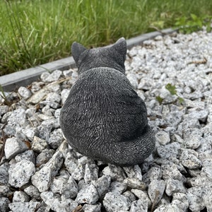 Puede incluir: Una figurita de gato de hormig&oacute;n gris sentada en un lecho de grava blanca.