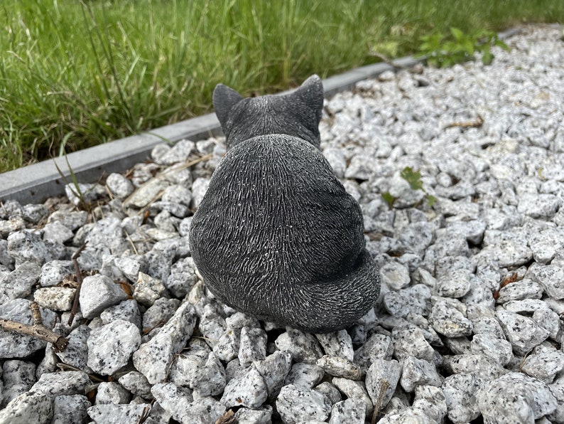 Puede incluir: Una figurita de gato de hormig&oacute;n gris sentada en un lecho de grava blanca.