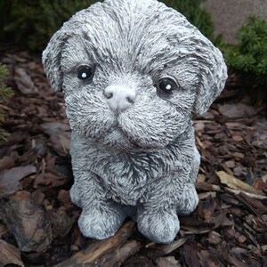 May include: A small, gray, furry, concrete dog statue. The statue is sitting on a bed of brown wood chips.