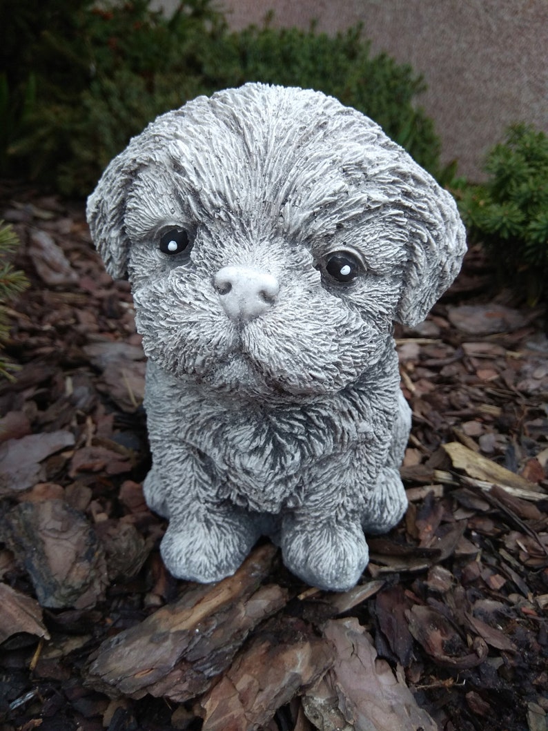 May include: A small, gray, furry, concrete dog statue. The statue is sitting on a bed of brown wood chips.