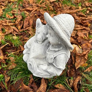 May include: A gray concrete garden statue depicting a child in overalls and a hat, reading a book with a dog. The statue is set on a bed of green grass and fallen brown leaves.