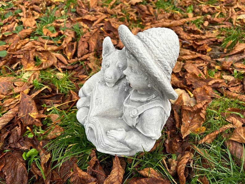 May include: A gray concrete garden statue depicting a child in overalls and a hat, reading a book with a dog. The statue is set on a bed of green grass and fallen brown leaves.