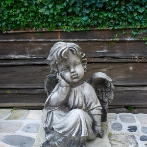 May include: A gray stone cherub statue with wings, sitting with its head resting on its hand. The statue is on a stone base and in front of a wooden wall with green ivy.