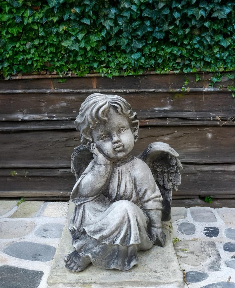 May include: A gray stone cherub statue with wings, sitting with its head resting on its hand. The statue is on a stone base and in front of a wooden wall with green ivy.