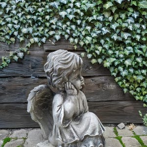 May include: A stone sculpture of an angel with wings, sitting with its head resting on its hand. The angel is wearing a flowing robe and is positioned on a stone base. The sculpture is set against a backdrop of a wooden wall covered in green ivy.