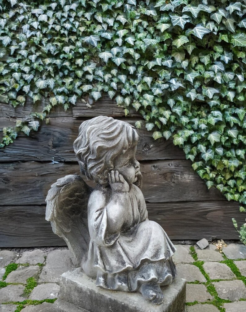 May include: A stone sculpture of an angel with wings, sitting with its head resting on its hand. The angel is wearing a flowing robe and is positioned on a stone base. The sculpture is set against a backdrop of a wooden wall covered in green ivy.