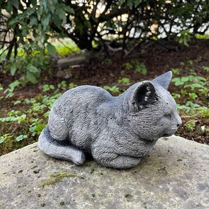 Puede incluir: Una estatua de gato de hormig&oacute;n gris en una pose de dormir. La estatua est&aacute; detallada con textura de pelaje y tiene una apariencia realista.
