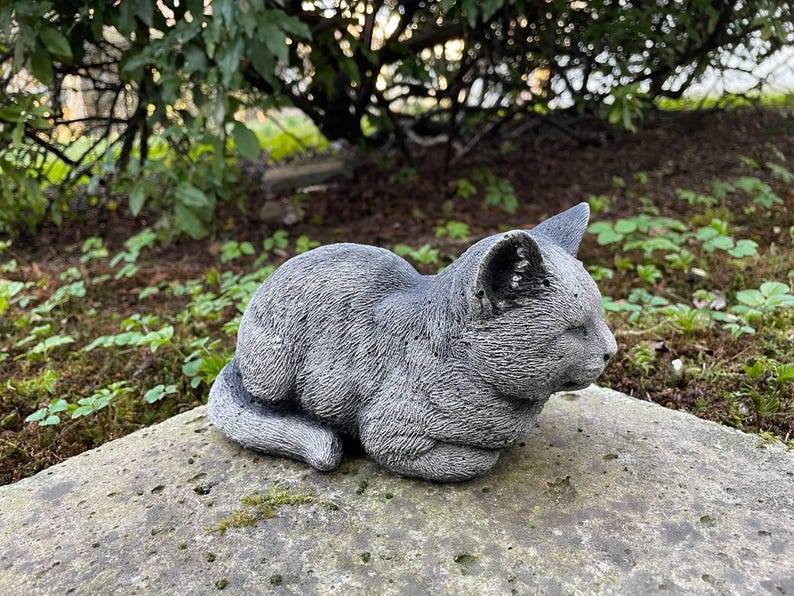 Puede incluir: Una estatua de gato de hormig&oacute;n gris en una pose de dormir. La estatua est&aacute; detallada con textura de pelaje y tiene una apariencia realista.