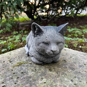 Puede incluir: Una escultura de hormig&oacute;n gris de un gato durmiendo con los ojos cerrados. La escultura est&aacute; sentada sobre una superficie de piedra gris.