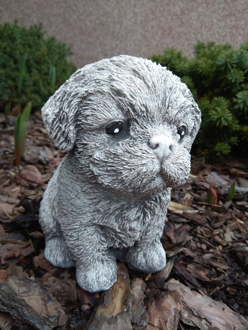May include: A small, gray, concrete statue of a puppy sitting on a bed of wood chips.