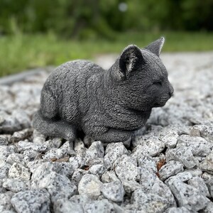 Puede incluir: Una estatua de gato de hormig&oacute;n gris con una expresi&oacute;n dormida, sentada en un lecho de peque&ntilde;as piedras grises.