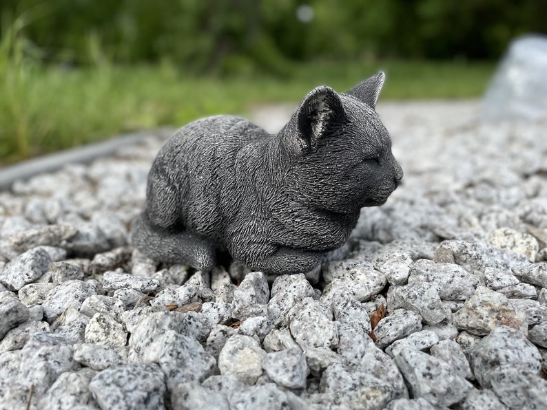 Puede incluir: Una estatua de gato de hormig&oacute;n gris con una expresi&oacute;n dormida, sentada en un lecho de peque&ntilde;as piedras grises.