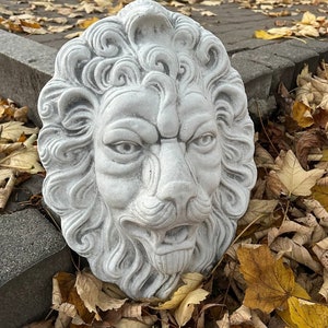 May include: A white concrete lion head sculpture with detailed features, including a mane, whiskers, and a snarling expression. The sculpture is resting on a bed of fallen autumn leaves.