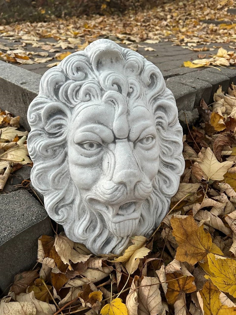 May include: A white concrete lion head sculpture with detailed features, including a mane, whiskers, and a snarling expression. The sculpture is resting on a bed of fallen autumn leaves.