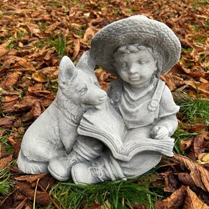 May include: A gray concrete garden statue depicting a young child wearing overalls and a straw hat, reading a book with a dog. The statue is set on a bed of brown leaves and green grass.
