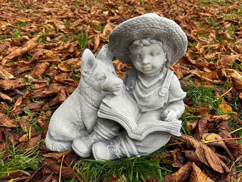 May include: A gray concrete garden statue depicting a young child wearing overalls and a straw hat, reading a book with a dog. The statue is set on a bed of brown leaves and green grass.