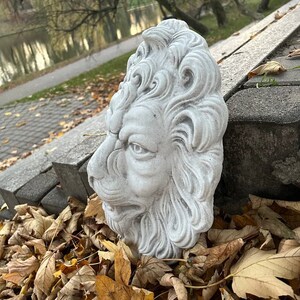 May include: A gray stone lion head sculpture with detailed carvings. The sculpture is placed on a pile of brown leaves.